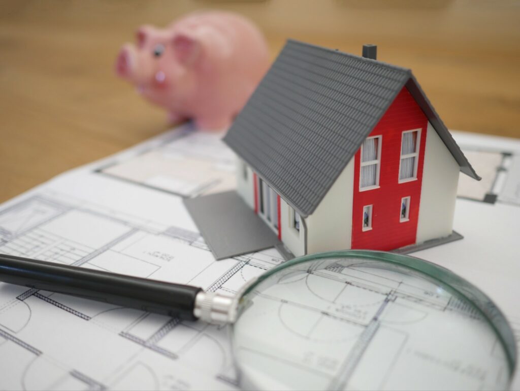 A piggy bank, A small to invest model of a house, and a magnifying glass on top of a floor plan.